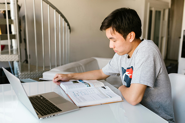 Asian man sitting a desk looking and book with a laptop computer in from of him